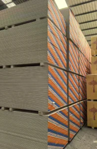 Large stack of drywall panels and orange-striped drywall in a warehouse setting, arranged neatly on pallets.