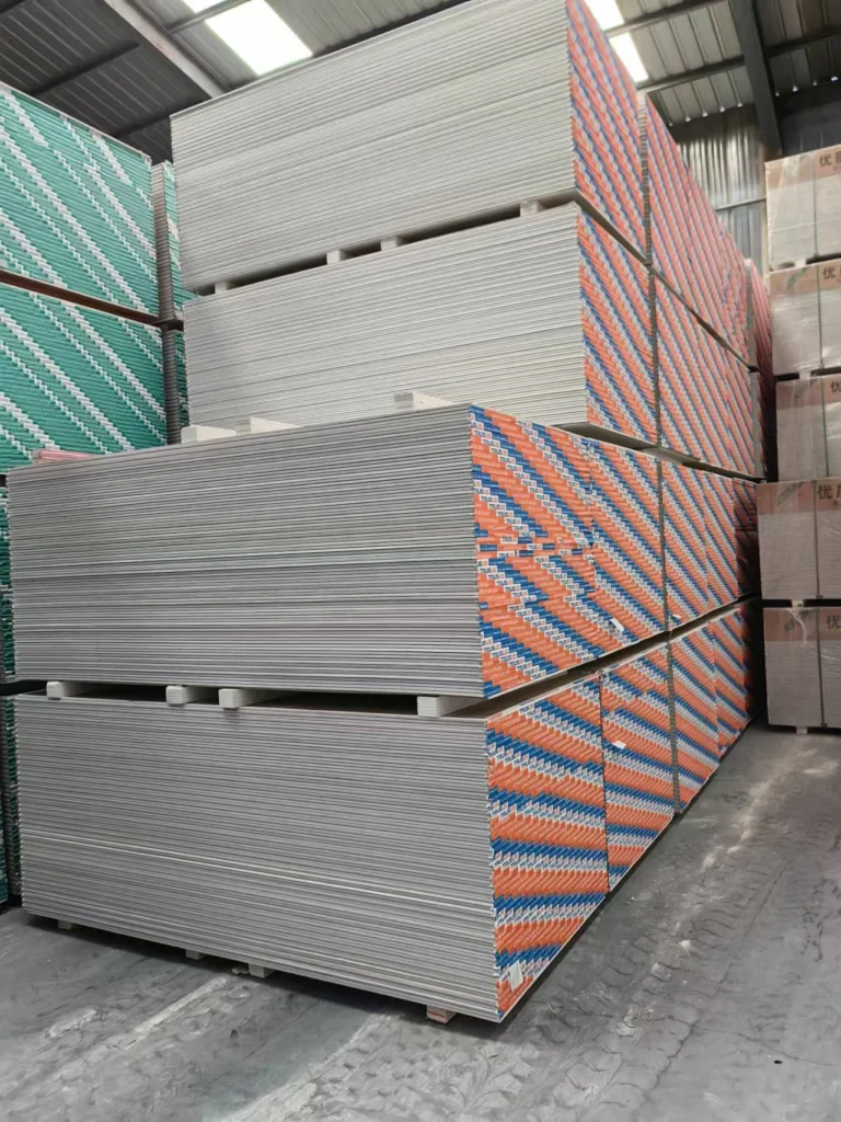 Stacks of drywall panels with colorful packaging in a large warehouse setting, indicating construction materials storage.