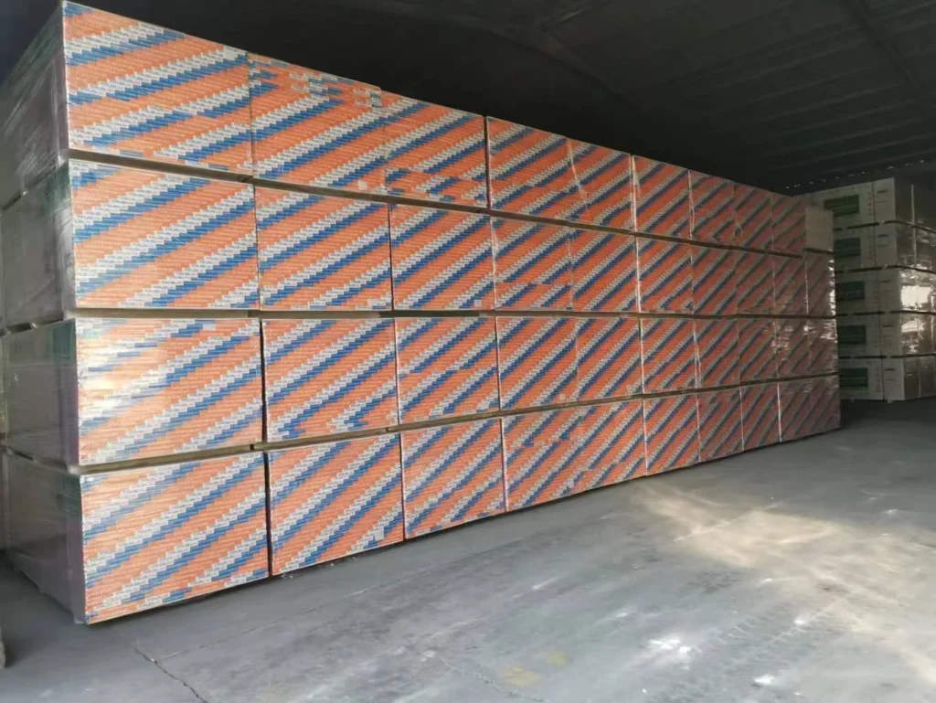 Large stack of drywall panels with orange and blue striped packaging in a warehouse setting.