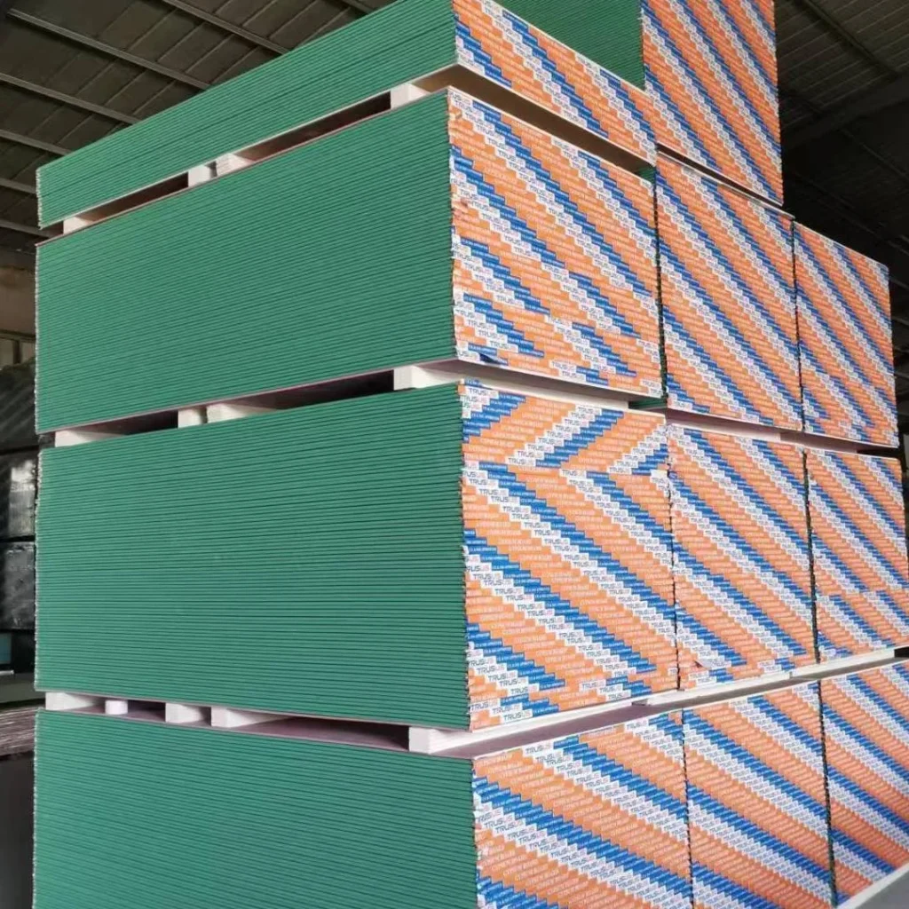 Large stack of green drywall panels stored in a warehouse along with panels wrapped in blue and orange packaging.